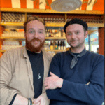 Two men standing side by side looking at camera. They are standing inside Finch Wine Bar with and orange and gold themed interior.