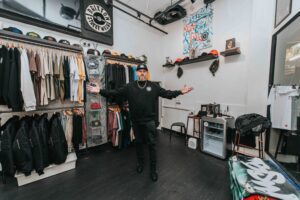 Dan standing inside he Brixton Street Wear store with his arms extended wide, smiling to camera.