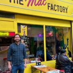 Man standing outside bright yellow storefront that reads 'The Mac Factory'