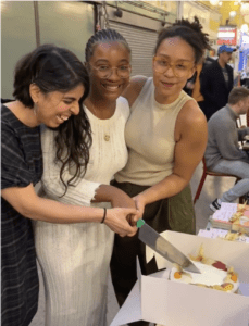 Three women holding a knife collectively cutting into a cake.