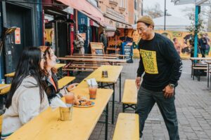 Two girls dining in the outdoor seating area of Fish Wings and Tings, Brian Danclair walking over to talk to them.